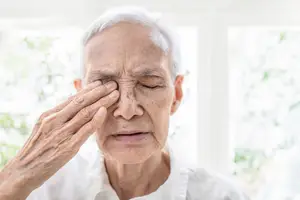 An elderly woman sitting inside the house with her eyes closed and hand on her forehead