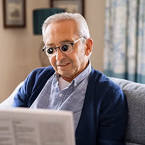 An elderly man with white hair and glasses is sitting on a couch and reading a newspaper.