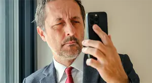 A middle aged man in a suit squints at his phone while standing in front of a window.
