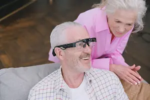 An elderly man and woman sit together on a couch, with the man wearing glasses with a built-in camera.