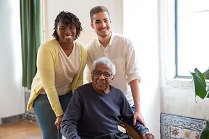 Three people smiling and standing in a room, a man sitting on a chair, and a plant on the right.