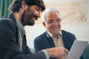 An older man and a younger man are sitting on a couch. The older man is wearing glasses and is smiling. The younger man is holding a tablet and is looking at the older man. Behind them is a wall with a decorative pattern. The older man is wearing a blue cardigan over a white shirt.