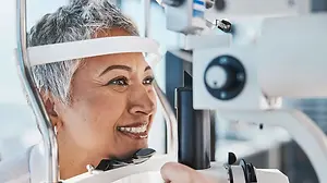 An older woman with short gray hair is smiling and looking through an eye examination machine with a white headband
