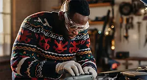 Man working on a car in a garage wearing a festive sweater, glasses, and gloves
