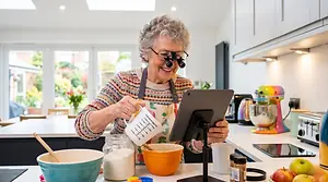 An elderly woman uses a magnifying glass to read a recipe on her tablet in the kitchen.