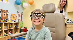 A young boy wearing eye test glasses in a doctor's office with a female doctor smiling in the background.