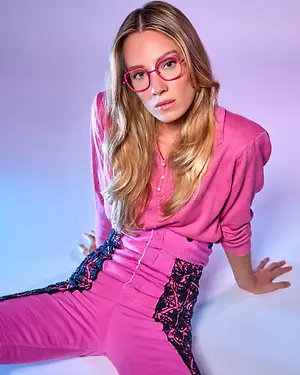 a woman wearing a pink blouse with a black lace design on the bottom is sitting on a white surface while posing for a photo