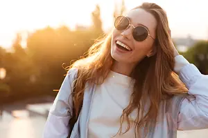 A smiling woman with long blonde hair wearing sunglasses and a white jacket standing in a park.