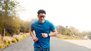 A man in a blue shirt and sunglasses is running on a road with trees and mountains in the background.