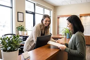A smiling woman is standing behind a reception desk, holding a clipboard and interacting with another woman in a green sweater, with a computer monitor and potted plants in the background.
