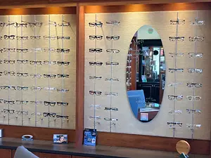 Inside a shop, there is a large mirror with a reflection of the shop's interior. The mirror is oval-shaped and has a frame, and it reflects the shop's interior, including a person and a desk. In front of the mirror, there is a display of eyeglasses, which are hanging on a wooden wall. The eyeglasses are arranged in rows and columns, and each pair has a different color and design. The shop has a wooden floor and a beige wall, and there are also other items on the wall, including a poster and a book.