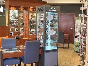 An empty optical shop with display glasses, a desk with chairs, a brown door, and a wooden cabinet with a glass display case with Oakley glasses and other items on it
