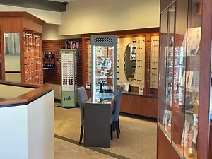 Inside an eyeglasses store, there is a display case with glasses on top of a desk, and two chairs are placed in front of the desk. On the left side of the store, there are shelves with different sizes of glasses. On the right side, there are mirrors mounted on the wall, and a round mirror is on top of the display case. On the back, there is a wooden shelf with a poster on it.