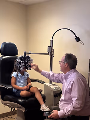 A man adjusts a child's eye exam equipment while holding a remote control.