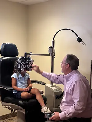 A man adjusts a child's eye exam equipment while holding a remote control.
