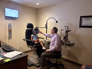 An adult male optometrist is examining a young girl's eyes using an ophthalmoscope in an office room.