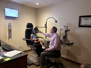 An adult male optometrist is examining a young girl's eyes using an ophthalmoscope in an office room.