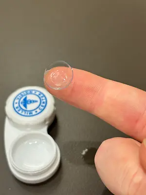 A finger holds a clear lens with a lens case behind it on a table