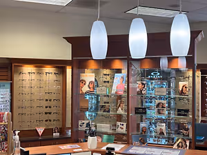 A photo of a store with glass cabinets displaying various eyewear, books, and a table with a white vase and other items. Three lamps are hanging from the ceiling.