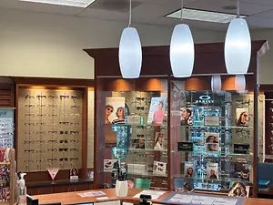 A photo of a store with glass cabinets displaying various eyewear, books, and a table with a white vase and other items. Three lamps are hanging from the ceiling.