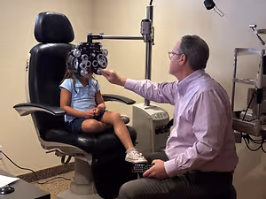 A man is examining a young girl's eyes using an ophthalmoscope in an eye care clinic.