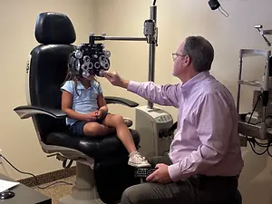 A man is examining a young girl's eyes using an ophthalmoscope in an eye care clinic.