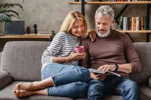 An older man and woman sitting on a couch with a book and coffee cup