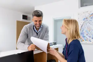 A man smiles while holding a piece of paper and talking to a woman in a blue uniform in an office setting.
