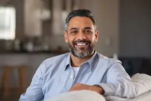 Smiling man with a beard wearing a light blue shirt sitting on a couch in a home interior.