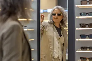 A woman is standing in front of a mirror and looking at her reflection. She is wearing sunglasses and a beige blazer. She is pointing to her face. Behind her is a rack of sunglasses. The rack has a gray wall with a black border and several shelves with sunglasses on them. The sunglasses are in different colors and styles.