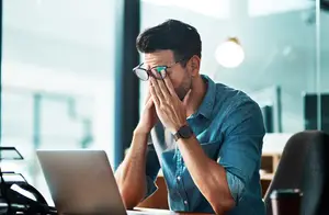 Stressed man in an office covering his mouth with his hand while sitting in front of a laptop