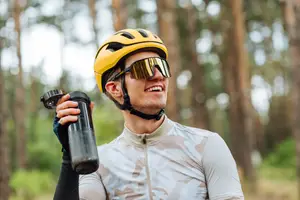 A man with a yellow helmet and sunglasses is smiling and holding a black water bottle in a forest.