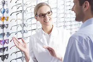 A woman with glasses stands in front of a man in a white shirt and talks to him while pointing to a wall with glasses in front of her