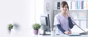 A woman dressed in scrubs is sitting at a desk and talking on the phone