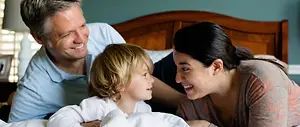 A family of three is smiling and looking at each other in a bedroom
