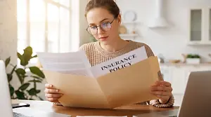 A woman wearing glasses is sitting in her home and reading an insurance policy document.