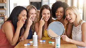 Five women look in a mirror and apply contact lenses, with one pointing to her eyes