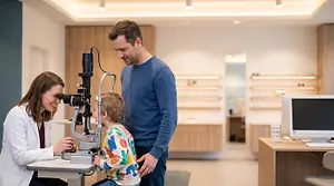 A man and a woman stand behind a child who is sitting in front of an optometrist examining machine while the optometrist holds the child's chin