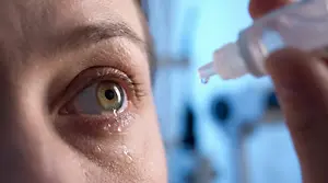 A person administering eye drops with one eye closed and teary, using a transparent bottle against a blue background