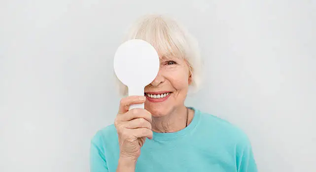 An elderly woman is holding a white mirror in front of her face and smiling