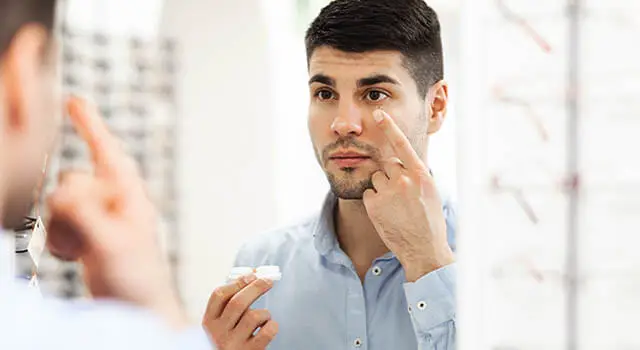A man looking at his eyes in a mirror while applying eye drops.