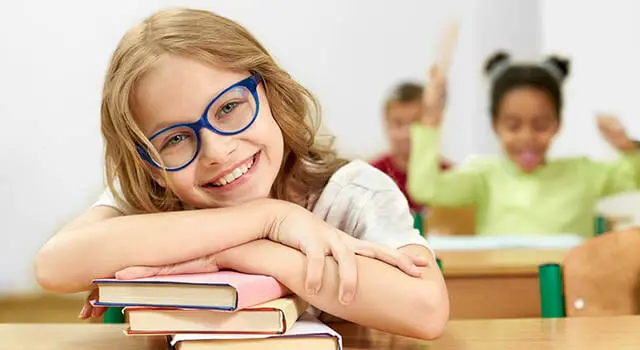 A young girl with glasses smiles in a classroom surrounded by other students.