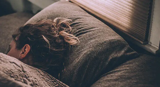 A woman with dark hair is sleeping on a couch under a window with blinds in a room. She has a pillow under her head and a blanket covering her body. She is wearing glasses and a dark hair tie.