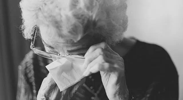An old woman wearing glasses is wiping her tears with a tissue while looking at something in front of her