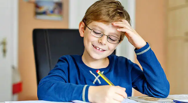 A young boy with glasses is smiling and holding a pencil while sitting at his desk